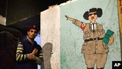 Сүрөттө: Муаммар Каддафинин Адолф Гитлердин кебетесинде тартылган карикатурасынын жанынан бир көтөрүлүшчү өтүп баратат.  Каддафи сол колуна "Менин Жашыл Китебим" деп аталган китепти кармап, оң колу менен нацисттик салам жолдоодо. Бенгази шаары, Ливия. 16.