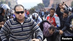 Эгіпецкі паліцыянт у цывільным бяжыць, каб напасьці на журналіста, які здымае зьбіцьцё пратэстоўцаў на заднім пляне.